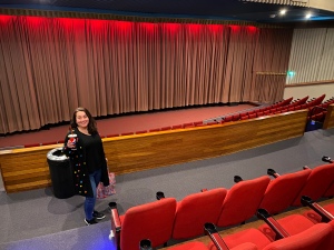 Natasha stands in an empty gala cinema with rows of red seats and the curtain drawn over the screen.