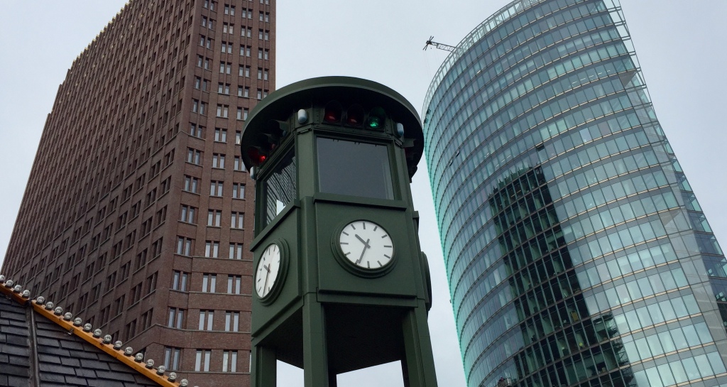 A clock on a watchtower with two buildings in the background and a market roof in the foreground, found in Potsdamer Platz, Berlin